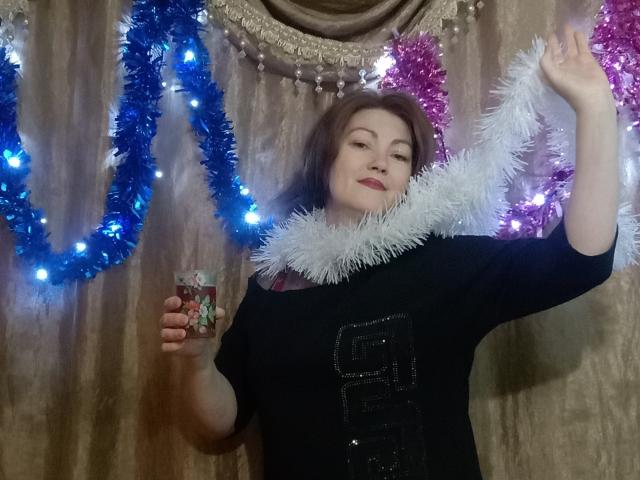 SadieLorri, con cabello castaño, vestida con una camisa negra con brillo, sosteniendo un regalo, con guirnalda blanca alrededor del cuello, levantando el brazo, con guirnalda azul y rosa con luces en el fondo..