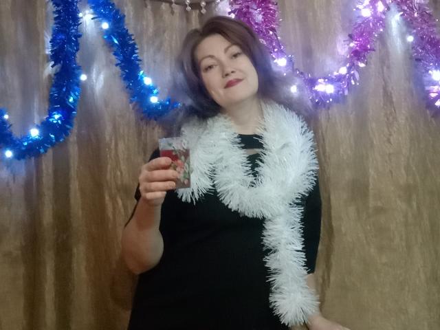 SadieLorri with purple hair, black dress, white tinsel necklace, holding a drink, smiling, blue and purple tinsel with lights in the background.