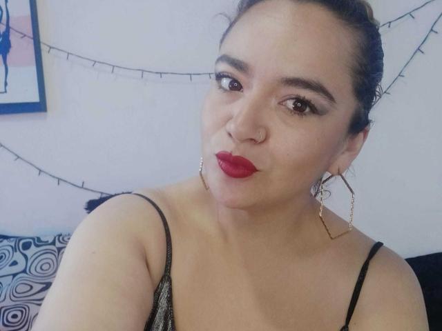 NaugthyCurvy, avec ses cheveux noirs attachés en chignon, son rouge à lèvres rouge, ses boucles d'oreilles cerceaux et son piercing au nez, porte un haut noir à bretelles fines qui dévoile sa poitrine et ses lèvres pulpeuses..