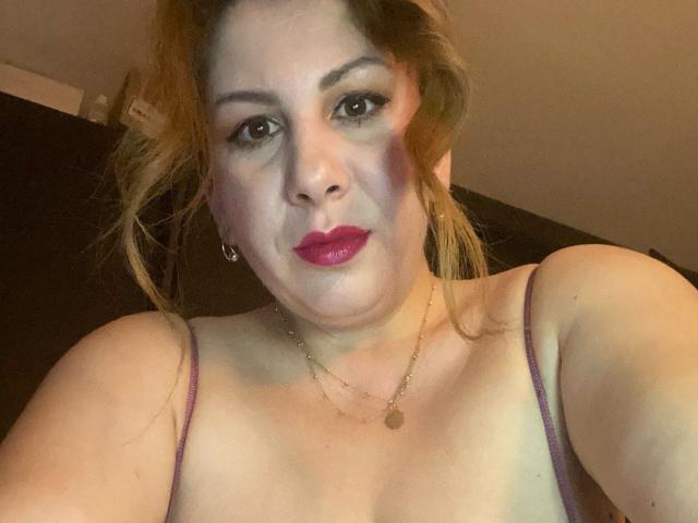 JolieJuliee com batom vermelho, vestindo uma blusa roxa de alças finas, mostrando os seios, cabelo loiro em um coque desarrumado, colar de ouro e uma expressão séria..