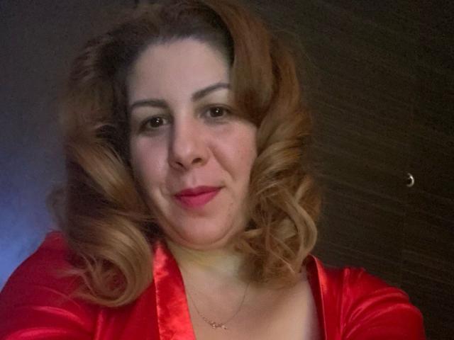 JolieJuliee, avec ses cheveux bruns bouclés, sa robe en satin rouge, son rouge à lèvres rose, son léger sourire et son collier, regarde directement l'appareil photo. Ses seins sont visibles sous la robe. L'arrière-plan est sombre et flou..