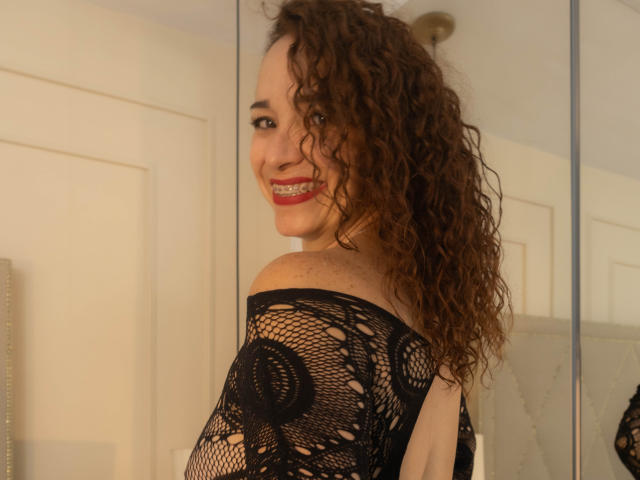 ScarlettthVega with curly brown hair, smiling, wearing a black fishnet top, showing her back and shoulders, red lipstick, white wall, mirror reflection, and a light-colored door.