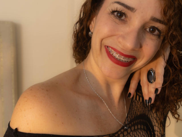 ScarlettthVega has curly brown hair, red lipstick, black nail polish, and a black lace top. She's smiling with a silver necklace and black ring. Her tits are slightly visible.
