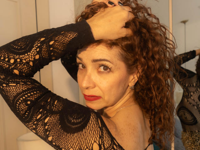 ScarlettthVega met krullend bruin haar, zwarte kanten mouwen, rode lippenstift en oorbellen, die haar haar omhoog houdt, kijkend naar de camera met een lichte frons, in een warm licht..
