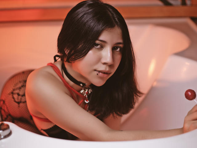 Emily Malhore, con cabello oscuro, vestida con una blusa roja y una malla de encaje negro, inclinada en una bañera blanca, sosteniendo un caramelo rojo, con un collar negro y un piercing en el labio..