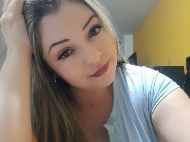 NatalyHami with long brown hair, dark eyeliner, and red lipstick, wearing a light blue shirt, smiling softly, hand in hair, looking at camera.