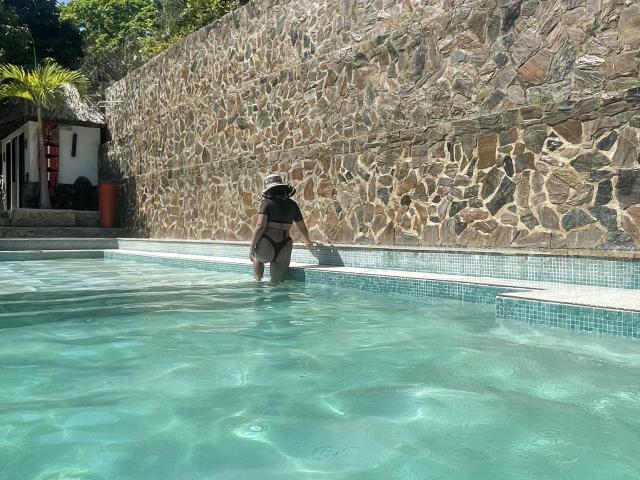 Il sedere di Zdiablaz è nella piscina, indossa un bikini nero e un cappello da sole, tiene un asciugamano, è in piedi vicino a un muro di pietra, con acqua cristallina intorno a lei..