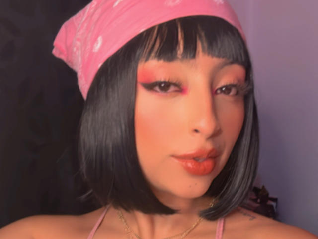 Zdiablaz trägt ein pinkes Bandana, einen schwarzen Bob, roten Eyeliner und glänzlich orangefarbene Lippen. Ihre Augen sind halb geschlossen, wodurch ihre langen Wimpern zur Geltung kommen. Sie trägt eine goldene Halskette..