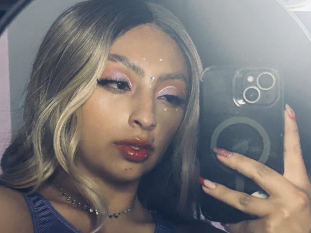 Zdiablaz has shiny blonde hair, pink eyeshadow, and glossy red lips, taking a selfie with a phone, showing a cute face with a small beauty mark on her chin.