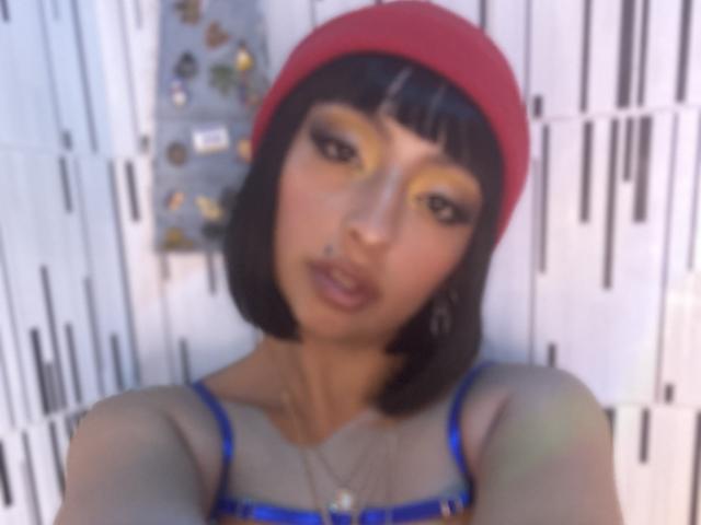 Zdiablaz ha un viso carino, un caschetto castano, un cappello rosso, ombretto giallo, un reggiseno blu e un piercing al naso, e guarda direttamente la fotocamera con un sorriso sottile e seducente..