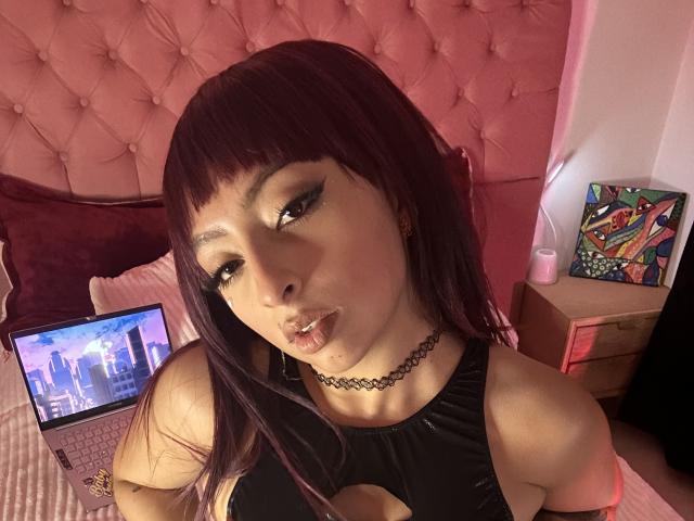 Zdiablaz, con cabello morado oscuro, un collar negro y una blusa negra con aberturas, labios fruncidos, una laptop mostrando un horizonte urbano en la cama con un cabecero rosa acolchado. Se ven los senos a través de la blusa..