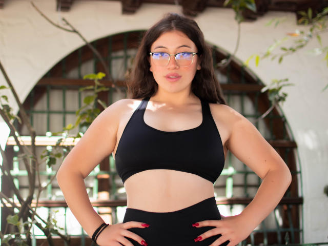 Amy Phillips, avec des lunettes, un soutien-gorge de sport noir et un pantalon noir assorti, les mains sur les hanches, des ongles rouges et un léger sourire, se tient là avec une allure confiante, sexy, pulpeuse et mignonne..