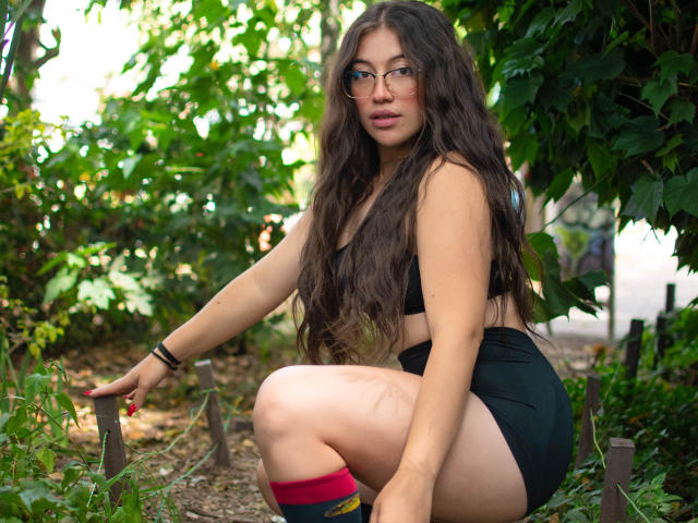 Amy Phillips, com cabelos castanhos longos, óculos, e um top e shorts esportivos pretos, ajoelhada em um jardim, mostrando sua nádega nua e meias vermelhas e pretas..