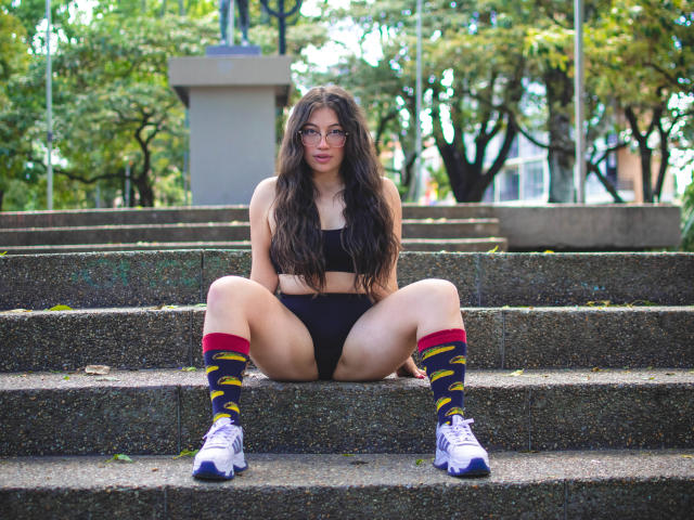Amy Phillips, com cabelos longos e escuros, óculos, um top de ginástica preto, calcinha preta, meias coloridas até o joelho e tênis brancos, sentada com as pernas abertas em degraus de concreto, expondo sua vulva..