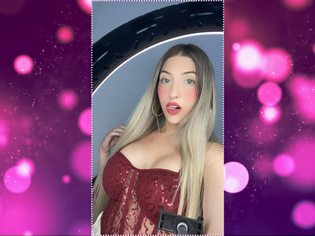 YejSusen com cabelos loiros longos, corpete de renda vermelha, seios grandes e brincos de argola dourados, segurando uma câmera, fundo rosa desfocado, fofa e sexy..