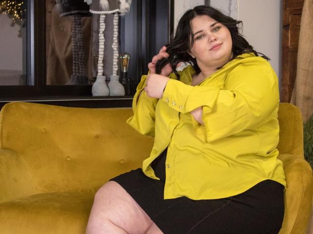 KaidoKun, une femme corpulente aux cheveux foncés, est assise sur un canapé jaune, vêtue d'une chemise jaune et d'une jupe noire, et elle touche ses cheveux avec un léger sourire..