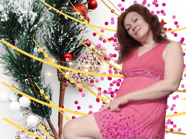 SadieLorri, met rood krullend haar, een roze jurk en zichtbare billen, staat bij een versierde kerstboom met gele linten, rode versieringen en roze kralen. Ze glimlacht, lichtelijk gekanteld..