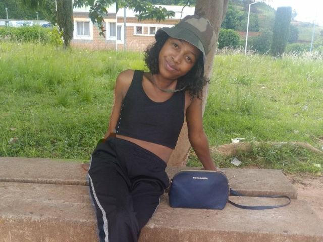QueenBella69 with dark skin, curly black hair, wearing a green bucket hat, black sports bra, and black track pants, sitting on a concrete bench, holding a blue bag.