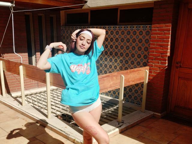 AnnaKhalo with pale skin, medium-length brown hair, wearing a turquoise shirt, white shorts, and a white headband, standing with arms raised, showing a tattoo on her left thigh.