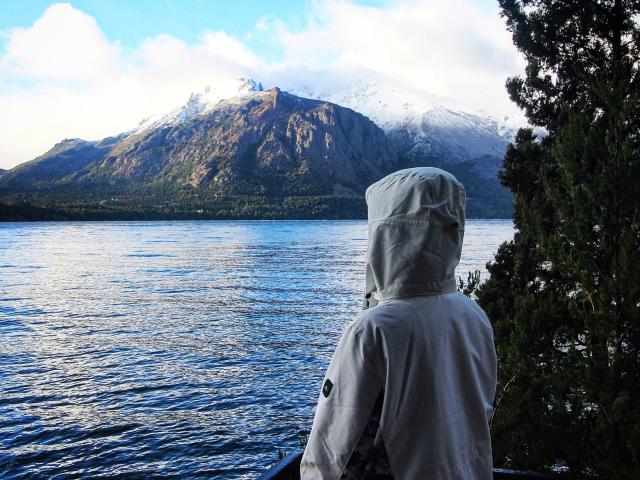 DelaMoons, con una sudadera gris y la capucha levantada, está de pie junto a un lago azul, frente a montañas cubiertas de nieve, rodeada de árboles verdes a la derecha..