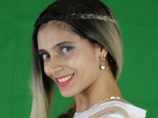 DelaMoons tem cabelo com efeito degradê de loiro para castanho, trançado, olhos verdes, batom rosa, vestido branco, brincos de pérola e um sorriso. Seus seios não estão visíveis..