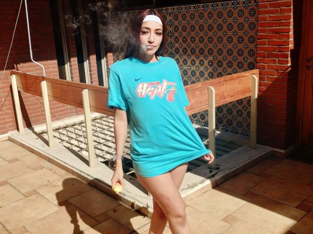 AnnaKhalo mit braunen Haaren, weißem Stirnband, blauem T-Shirt und ohne Hose, raucht, wobei ihr nackter Hintern sichtbar ist. Sie steht auf einer sonnendurchfluteten Ziegelterrasse, neben einem Holzzaun, und im Hintergrund ist ein Fliesenmuster zu sehen..