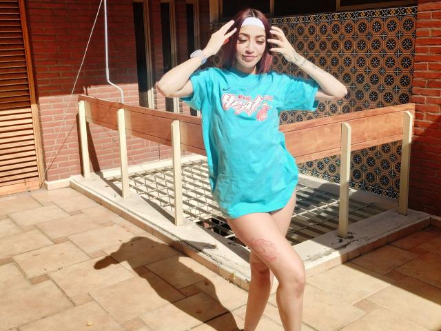 AnnaKhalo, à la peau pâle, aux cheveux roux, portant une chemise turquoise ample qui expose ses cuisses, avec un tatouage sur sa jambe droite, se tient dans une cour pavée éclairée par le soleil, les mains dans les cheveux..