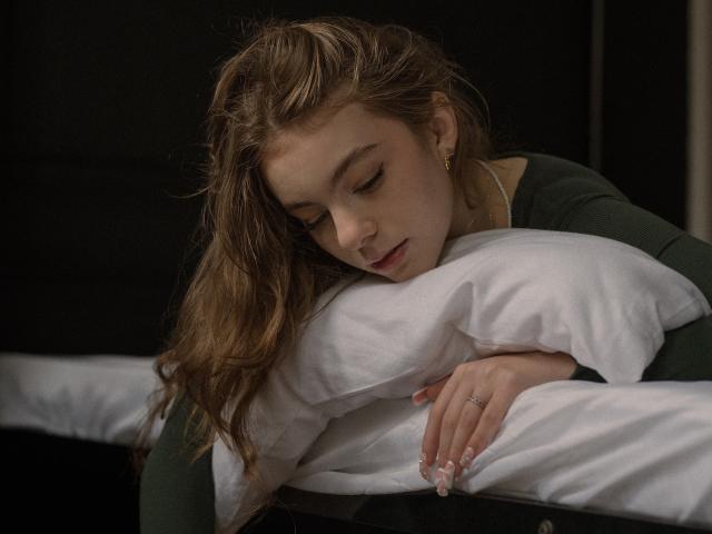 AriaMiss with wavy brown hair, green long-sleeve shirt, hugging a white pillow, eyes closed, soft expression, ring on finger, dark background, realistic, tender, peaceful.