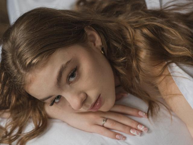 AriaMiss with wavy brown hair, green eyes, and a ring on her hand, lying on a white bed, looking up with a pouty expression. Her nails are manicured.