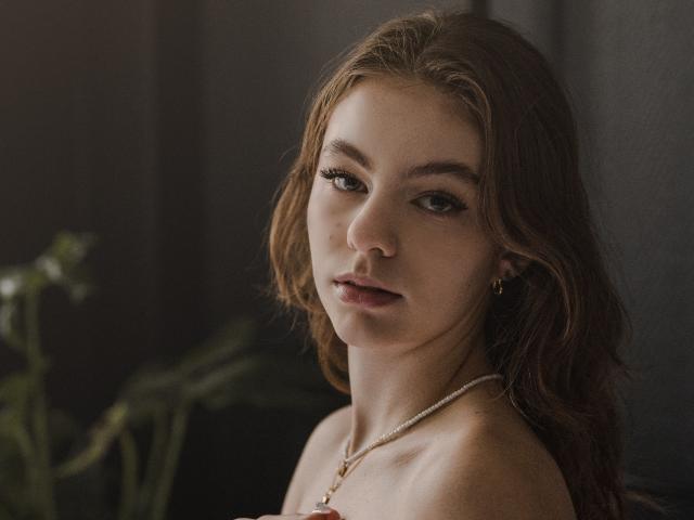 AriaMiss has wavy brown hair, light skin, and brown eyes. She's wearing a necklace with a pink gem, and small gold earrings, with a subtle, sexy look.
