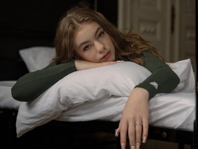 AriaMiss with long brown hair, green long-sleeve shirt, and white pillow, resting her head and arm on a bed, looking tired and slightly sad. Her hand has a ring.