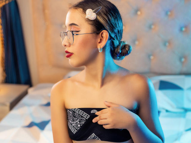 Valentina Cortes, avec des lunettes, du rouge à lèvres, et une robe noire sans bretelles, tenant ses petites seins, portant une pince à cheveux fleurie et des boucles d'oreilles en diamant, dans une pièce éclairée d'une douce lumière bleue..