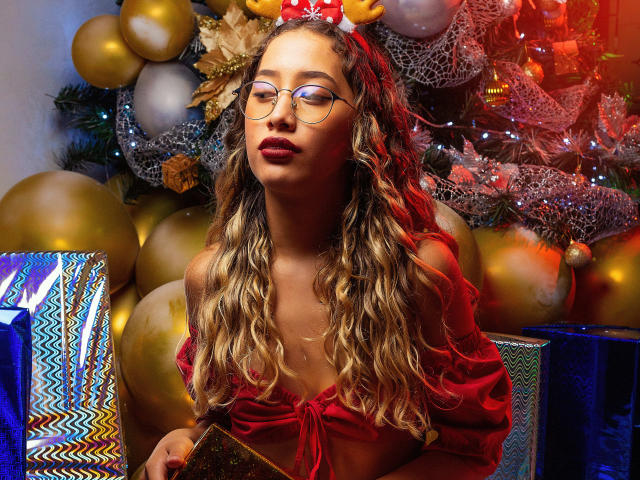 Valentina Cortes, com cabelos loiros ondulados, blusa vermelha com um ombro à mostra, óculos e uma tiara vermelha, segurando um presente, em frente a uma árvore de Natal decorada com balões dourados..