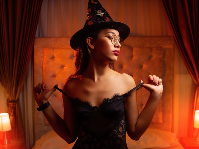 Valentina Cortes, usando óculos, um chapéu de bruxa e lingerie de renda preta, segurando as alças, com os pequenos seios visíveis, pele morena, batom vermelho, em pé em um quarto com pouca luz, olhando para o lado..