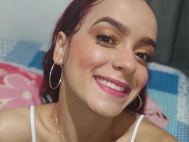 MaiaPark has red hair, a nose piercing, and large hoop earrings. She's smiling with pink lipstick, golden eyeshadow, and a white top. Her tits are slightly visible.