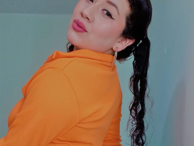 VeronicaHot69 met volmaakte, roze lippen, draagt een oranje geribbelde top, zwarte vlechtjes en grote oorbellen, en kijkt over haar schouder, waarbij haar rug zichtbaar is..