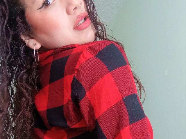 VeronicaHot69 con capelli castano ricci, camicia a quadri rossa e nera, bocca leggermente aperta, rossetto rosso e orecchino a cerchio argentato, che guarda oltre la spalla, con una parete verde sullo sfondo..