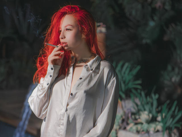 Camille Rios, com cabelo ruivo, fumando, vestindo uma camisa branca de botões, um colar e olhando para o lado. Ela tem uma estrutura física esguia e seios pequenos. O fundo está desfocado..