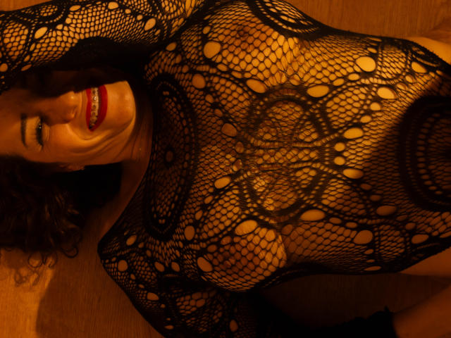 ScarlettthVega con lencería de malla negra, sonriendo con lápiz labial rojo, mostrando sus pechos y vulva a través de la tela estampada, acostada en un suelo de madera, con los brazos por encima de la cabeza..