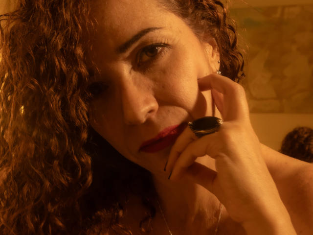 ScarlettthVega mit lockigem, braunem Haar, rotem Lippenstift, die einen schwarzen Ring in der Nähe ihres Kinns hält, verführerisch in die Kamera blickt, ihre Augen halb geöffnet, in warmem, goldenem Licht..