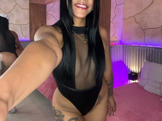 Amberv, con cabello negro largo, sonriendo, vestida con un traje de baño negro transparente que revela sus pequeños senos y una tanga negra, está de pie en un dormitorio con una cama rosa..