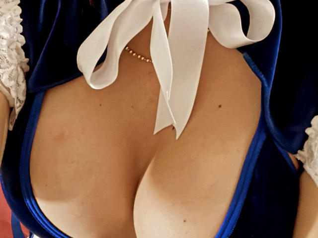 Os seios grandes e arredondados da ScarletBeautty em um vestido de veludo azul com mangas de renda branca e um laço branco amarrado no topo, exibindo um colar de ouro..