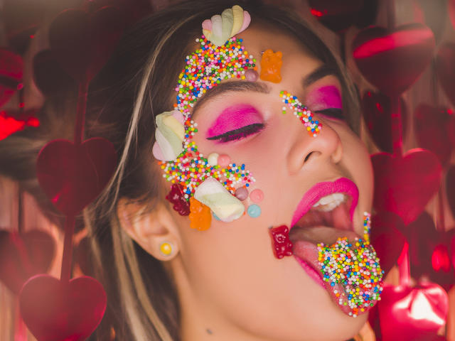 SunriseX's face covered in colorful candy, heart-shaped lollipops, and sprinkles. Pink eyeshadow, bright lipstick, tongue out, looking up. Red heart-shaped objects in the background.