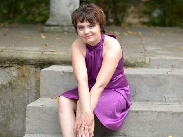 Kitti, avec ses courts cheveux bruns, sa robe en satin violet, assise sur des marches en béton, les mains posées sur ses genoux, souriant légèrement, peau pâle, mignonne, petite poitrine, maquillage discret, cadre extérieur décontracté..