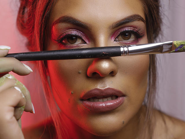 SunriseX's face with dark brown eyes, pinkish-purple eyeshadow, and glossy pink lips. She's holding a paintbrush in front of her nose, showing small skin blemishes. Red light.