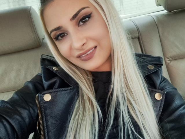 BonnieBlossom, met blond haar, draagt een zwarte leren jas, glimlacht, toont haar parelwitte tanden en ziet er schattig uit met haar make-up, waarbij haar borsten licht zichtbaar zijn..