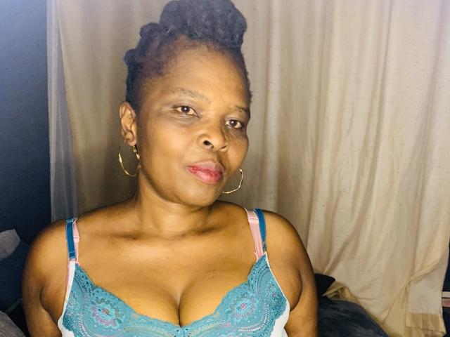 SexyRubi69 com pele marrom escura, cabelo cacheado preso em um coque, usando um sutiã de renda azul que mostra seus seios grandes, brincos de argola dourados e alças rosa, olhando ligeiramente para o lado..