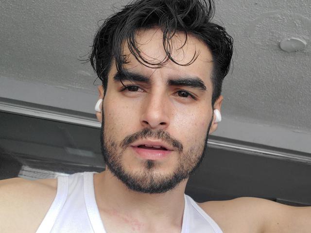 MateoGrand with dark, messy hair, white earbuds, slight beard, and white tank top, has a neutral expression and slightly parted lips, showing his nose and upper chest.