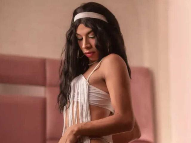 SalomeGrey with long black hair, wearing a white headband and sheer, fringed top, exposing her small tits and side of her ass. Soft pink couch in background.