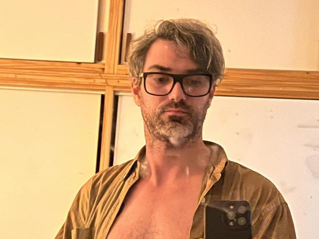 Kaik has gray, messy hair, a beard, and wears black glasses. He's shirtless with a brown, unbuttoned shirt, showing chest hair, and holds a black phone.